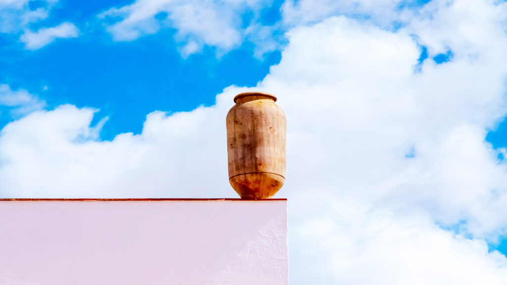 Tinaja tradicional de cerámica colocada como elemento decorativo en la esquina superior del edificio, símbolo del Club Deportivo y Cultural "Las Tinajas", destacando contra un cielo azul con nubes.