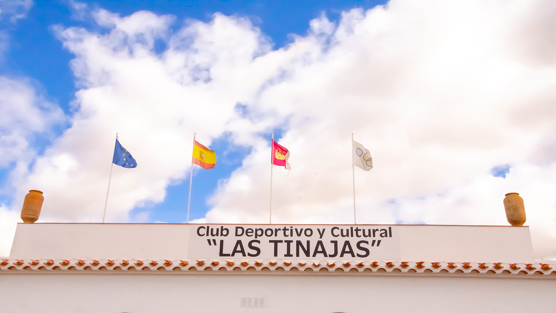 Cartel del Club Deportivo y Cultural Las Tinajas con banderas ondeando sobre fondo de cielo con nubes