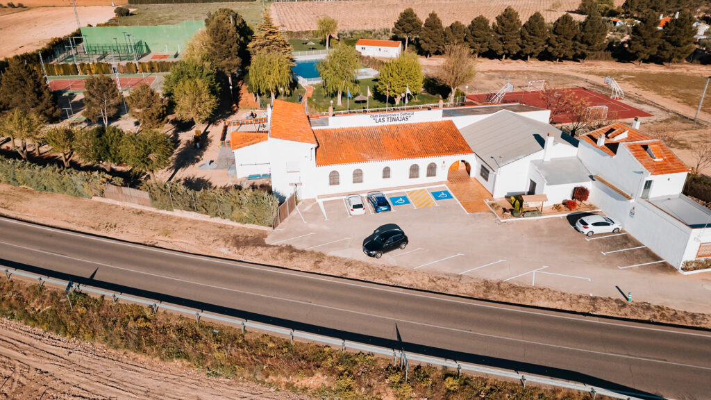 Vista frontal del edificio del club con pistas deportivas, piscina y zonas verdes al fondo.