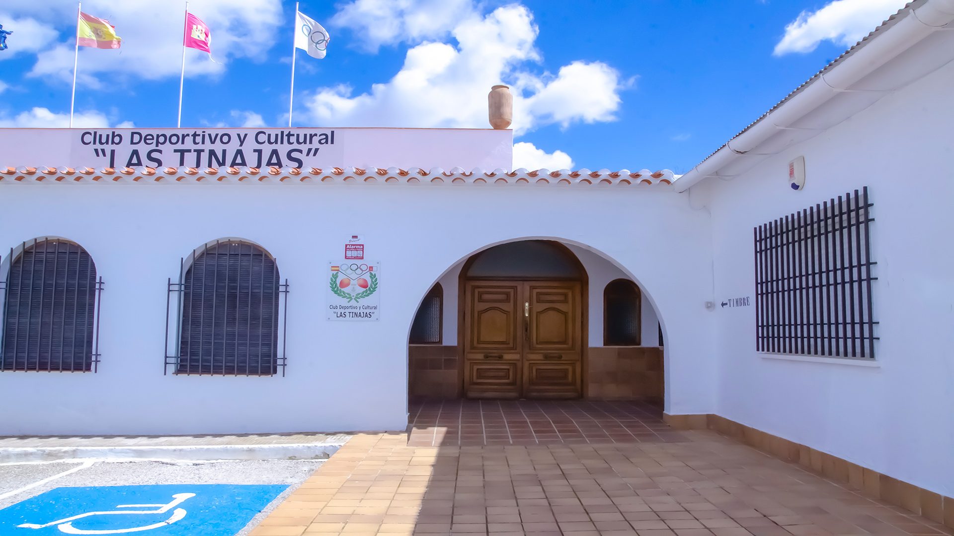 Entrada principal Club Las Tinajas Entrada principal del Club Deportivo y Cultural "Las Tinajas", con fachada blanca, puerta de madera, acceso adaptado para personas con movilidad reducida y banderas ondeando bajo un cielo azul con nubes.