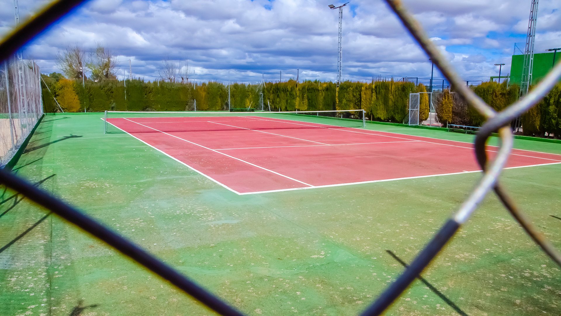 Pista de tenis Club Las Tinajas vista exterio Pista de tenis al aire libre del Club Deportivo y Cultural "Las Tinajas", con superficie roja y verde, rodeada de setos y protegida por malla metálica, vista a través del enrejado perimetral.