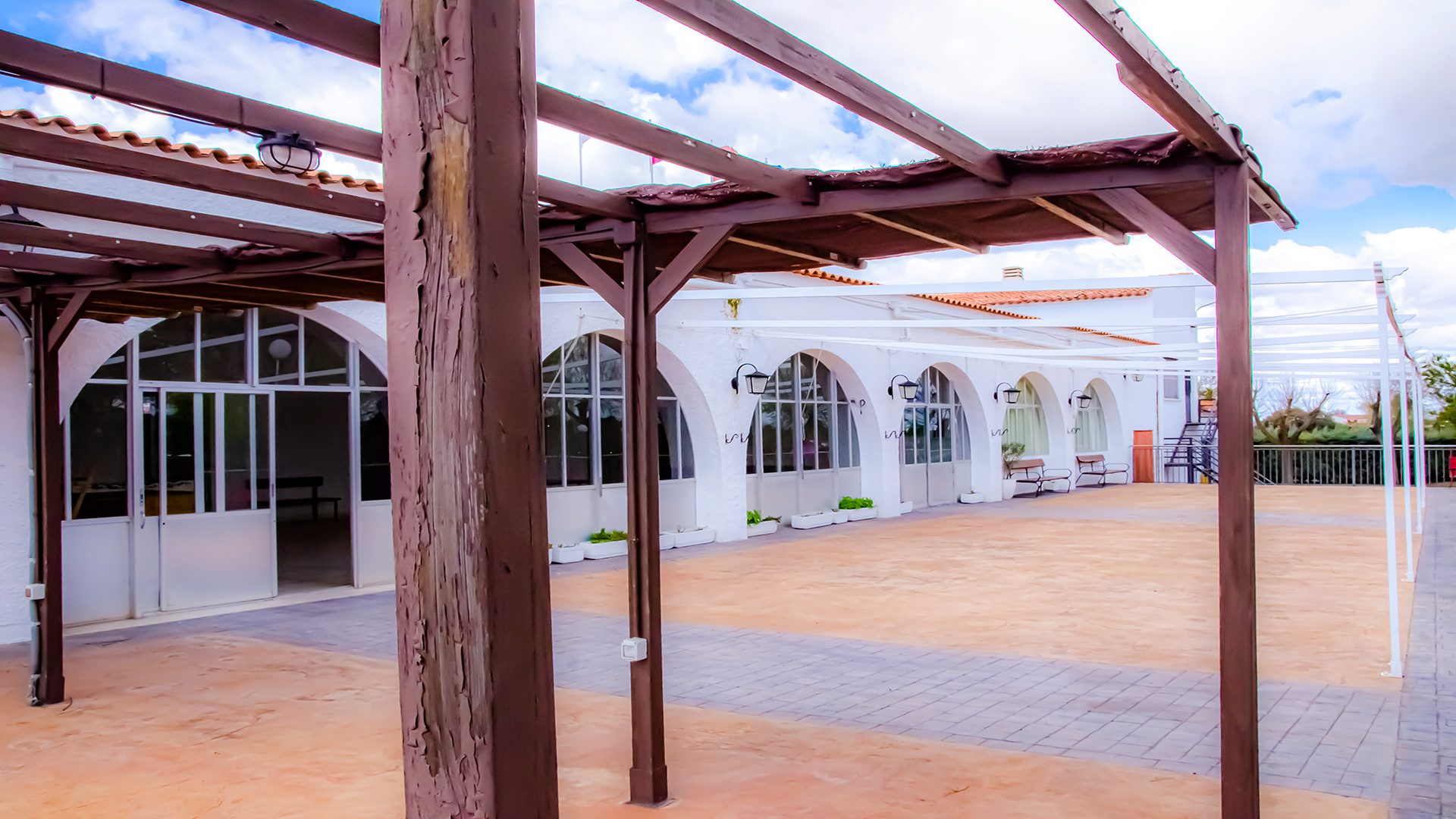 Vista de la amplia zona exterior de terraza y restauración del Club Deportivo y Cultural "Las Tinajas", con pérgola de madera, amplios ventanales arqueados y espacio abierto para eventos.