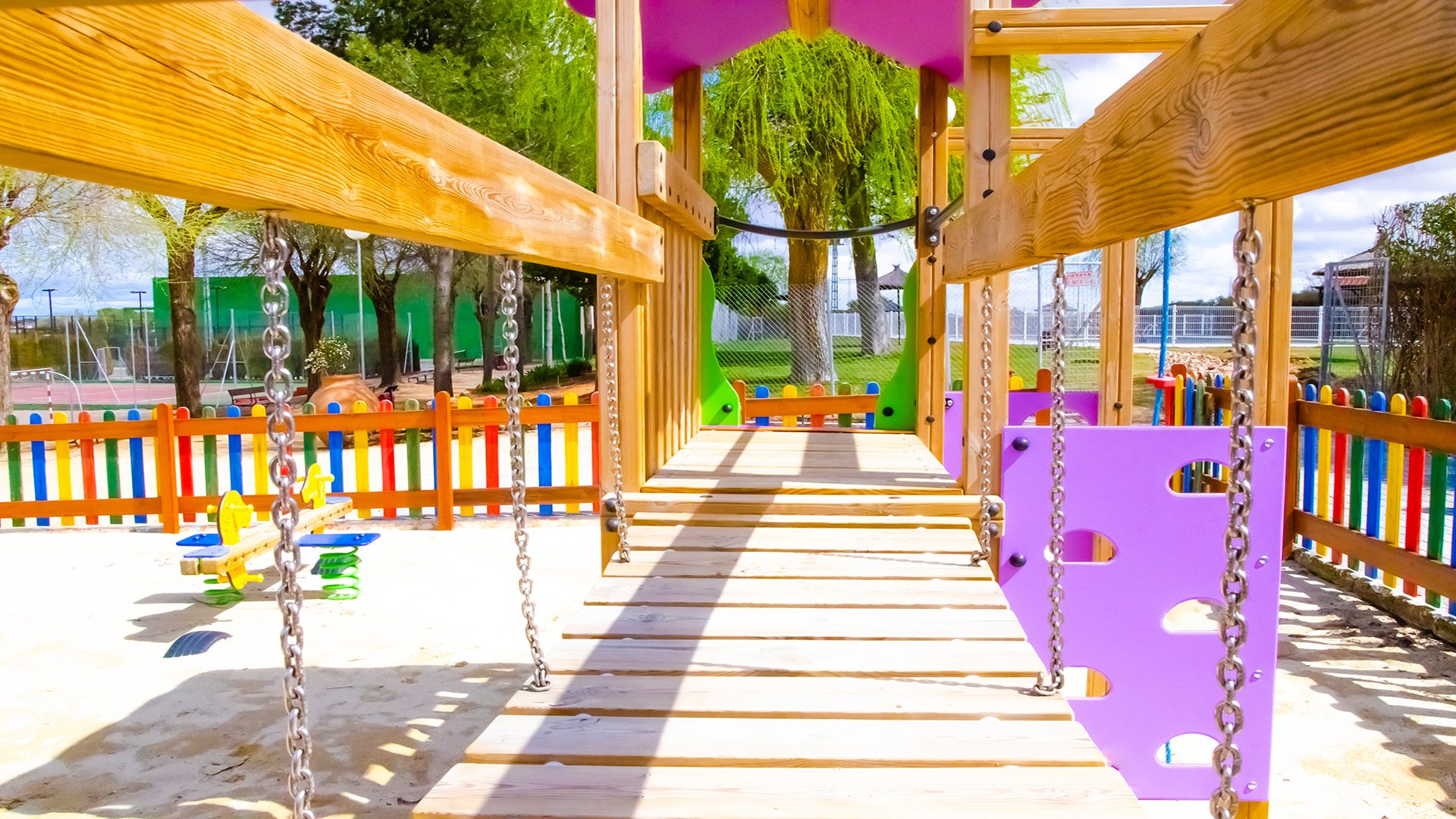Zona infantil del Club Deportivo y Cultural "Las Tinajas", con estructura de juegos de madera y cadenas metálicas, elementos coloridos y vallas de madera multicolor, en un entorno seguro y soleado.